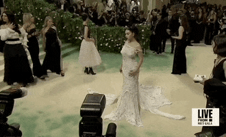 Met Gala 2024 gif. Charli XCX poses with hands on her hips wearing a white Marni gown with hand-sewed rhinestones and glass beads made of ripped patchworked well-worn t-shirts with holes throughout. The top of the off-the-shoulder dress resembles a ripped and worn t-shirt neckline with large holes wrapping around the upper arms.