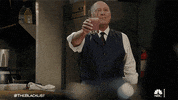 TV gif. James Spader as Red in The Blacklist. He is wearing a suit in a kitchen and he smiles and lifts a glass filled with a smoothie in cheers before bringing it to his lips to take a sip.