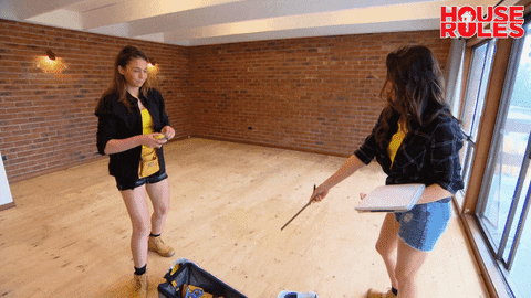 Reality TV gif. Two contestants on House Rules are measuring their home and one of them pulls a measuring tape out and holds it above her head.