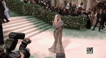 Met Gala 2024 gif. Kim Kardashian wearing a silver Maison Margiela gown and gray cardigan bolero, poses shuffles side to side, and waves into the distance.