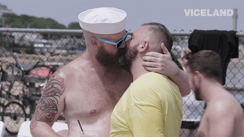 Video gif. Shirtless bearded man in a sailor hat makes out with another bearded man while people pass by behind them.