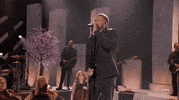 Academy of Country Music Awards gif. On a stage with instrumentalists and cherry blossoms in the background, Kane Brown sings into microphone intently. Brown's eyes start closed before slightly opening them when he finishs singing note with a pleased look