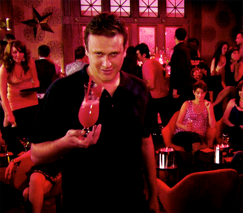 Movie gif. Jason Segal as Peter in Forgetting Sarah Marshall holds a piña colada awkwardly and sways, smiling creepy at us, in the foreground of a bustling party.