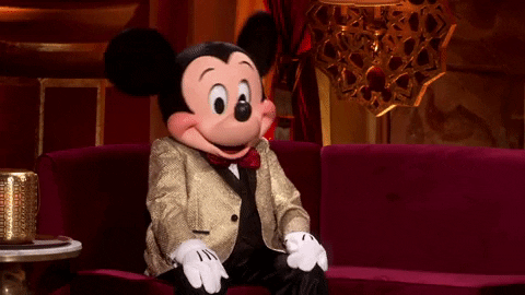 Disney gif. Mickey Mouse, sitting on a couch in an ABC studio dressed in a gold tuxedo, bobbing his head and shrugging, smile frozen on his face.