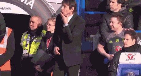 Sports gif. Antonio Conte cheers and jumps into a group hug in the stands at Stamford Bridge stadium.