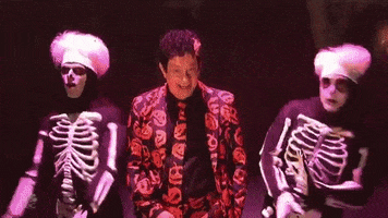 SNL gif. Tom Hanks as David S. Pumpkin on SNL, wearing a pumpkin-patterned suit and tie stands with a sinister smile as two other cast members dressed as skeletons dance, before Hanks smacks the skeleton’s backsides to get them to stop and pose.