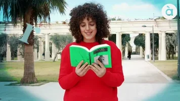 Mujer sonriente lee un libro verde en un parque, con un logotipo circular visible en la esquina superior derecha