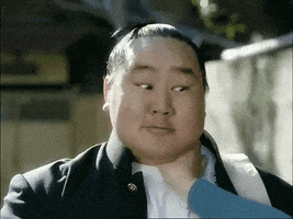 Video gif. Someone has an Asian man in a chokehold, but he gives them a thumbs up and blinks both eyes in a failed attempt to wink.