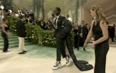 Met Gala 2024 gif. Alton Mason wearing a tweed ensemble and cape by Thom Browne, shuffles across the carpet confidently in towering platform boots, stopping to pop a leg and pose for the paparazzi.