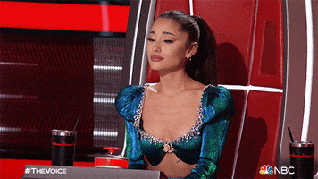 Reality TV gif. Performer Ariana Grande clasps her hands in front of her chest and fawns with admiration from her judge's seat on The Voice.