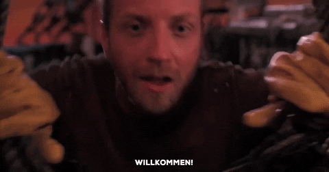 Movie gif. Chris Elliot as Nathaniel Mayweather from Cabin Boy leans over a drape and says enthusiastically, "Willkommen!," which appears as text.