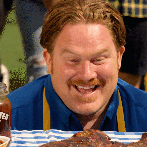 Celebrity gif. Casey Webb looks down at a table full of food with a giddy smile and excited eyes. Someone sides a plate full of burger sliders and his eyebrows lift in excitement.