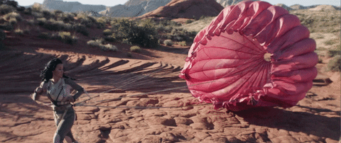 Music Video gif. Katy Perry in the Rise music video runs in the desert with a big pink parachute dragging behind her.  She has a hopeful expression on her face. 