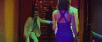 Music video gif. From the video for Neon Indian's Slumlord Rising, a man in a white suit stands at the foot of a sparkling staircase and waves his hand, welcoming a man and woman approaching the staircase.