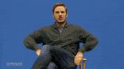 Celebrity gif. Chris Pratt sits atop a director's chair in front of a blue screen, which transforms into a wondrous, cosmic background as he theatrically raises his arms for us to behold the galaxy behind him.