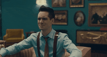Music video gif. In a clip from the video for Taylor Swift's "ME!", Brendon Urie wears suspenders and a tie over a powder blue shirt as he throws his hands up in an exasperated shrug. 
