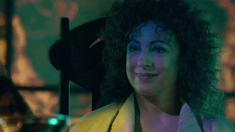 TV gif. Alex Kingston as River Song from Doctor Who smiles at us and raises a glass of champagne.
