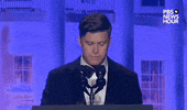 Video gif. Saturday Night Live's Colin Jost stands at a podium at the 2024 White House Correspondents' Dinner as he looks up at the crowd with a deadpan expression. He mischieviously looks to the left side of the room, then to the right, and looks down at his speech before smirking slightly.