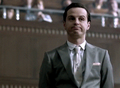TV gif. Andrew Scott as Moriarty on "Sherlock" in a courtroom giving an awkward straight-faced expression, which morphs into a frowning grimace as he turns around to look at Watson, played by Martin Freeman.