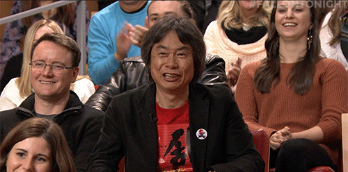 Celebrity gif. Shigeru Miyamoto, in TV audience, extends a thumbs down and frowns, but then turns it into a thumbs up and laughs as the crowd laughs and cheers around him.