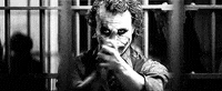 Movie gif. Black and white clip of Heath Ledger as The Joker in The Dark Knight. He stands in a jail cell clapping rhythmically as he glares down at us.