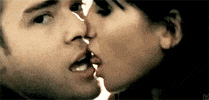 Music video gif. From Cry Me a River, Justin Timberlake looks and smiles at us while a dark-haired woman licks his face sensually.