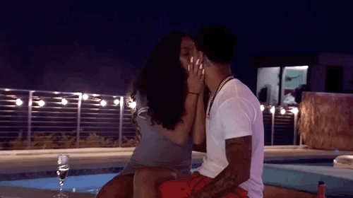 Reality TV gif. Sitting poolside at night in Ex on the Beach, a man and woman kiss.