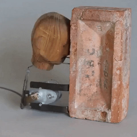 Video gif. Machine that bangs a wooden head against a brick repeatedly.