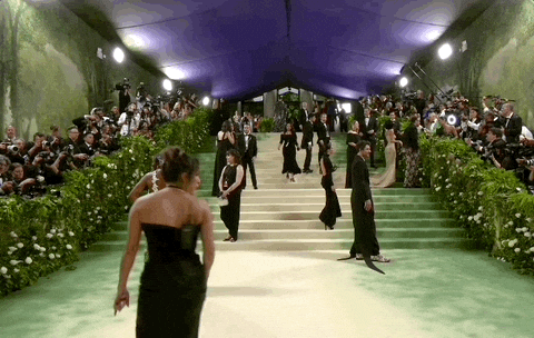 Met Gala 2024 gif. Wide shot of the carpet speckled with publicists, assistants, and celebrities as Mike Faist walks into the foreground photobombing the camera, sticking his tongue out and throwing us a peace sign as he passes through.