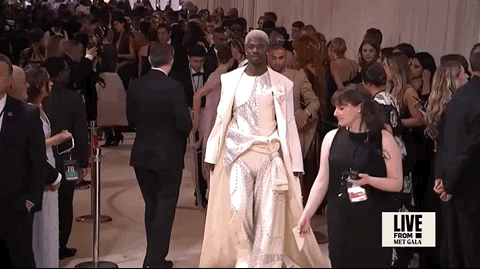 Met Gala 2024 gif. Lil Nas X struts down a hallway filled with people wearing an ivory Luar suit featuring asymmetrical panels of silver Swarovski sequined fabric with long lines of cream-colored buttons. His drapey wide leg pants billow out as he walks. His hair is dyed pale blonde 