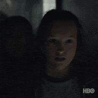 Video gif. Bella Ramsey as "Ellie" in the post-apocalyptic TV show "The Last of Us" is in a dimly lit corridor, looking in horror at something in the distance. The camera cuts to reveal a flashlight-lit, cordyceps-infected zombie in tattered clothing turning around to roar. 