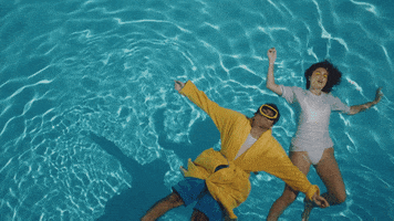 Music Video gif. From K.I.D.'s video for Errors, man floats on his back in a pool, wearing yellow goggles and a robe, next to a woman floating on her back, dressed in white.