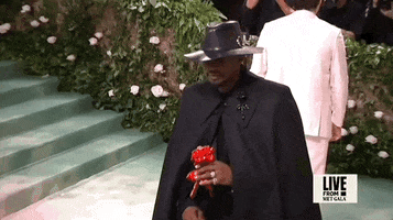 Met Gala 2024 gif. Usher, wearing an all-black custom Alexander McQueen ensemble, eyes spookily hidden by his fedora, holds a red paper flower.