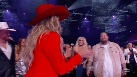 ACM Awards gif. Country music artist Lainey Wilson with a smile on her face claps while walking up like an old friend to Jelly Roll to give him a hug and as they seperate one of his hands linger on her forearm before he walks away completely. 