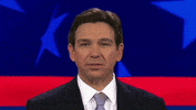 Video gif. Ron DeSantis gives a slight smile as he flicks his tongue out to touch his top and bottom teeth in front of a patriotic background. 
