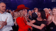 ACM Awards gif. Lainey Wilson and woman in black off-the-shoulder dress exchange a hug before the woman in black gives Lainey a peck on the cheek while bearing a congratulatory expression. 