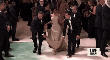 Met Gala 2024 gif. Tyla takes short steps in high heels as four assistants carry her train. She's wearing a sand-colored sculpted Balmain mermaid-style dress that's completely fitted through her angled waistline and hips. The fabric through the skirt is stiff and structured, giving the appearance of waxed fabric. 
