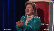 TV gif. On an episode of "The Voice," judge Kelly Clarkson claps and cheers joyfully from her judge's chair.