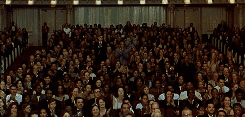 Movie gif. In a huge, fancy auditorium with multiple balconies, the entire audience begins to stand up and applaud.