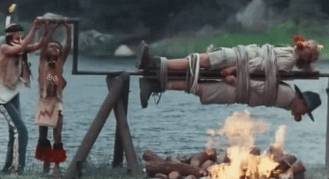 Movie gif. From Addams Family Values, two young campers dressed as Native Americans spin Peter MacNicol as Gary and Christine Baranski as Becky tied-up and gagged on a spit over a campfire.