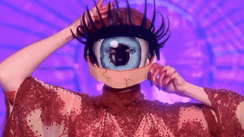 TV gif. An eerily beautiful model from Drag Race UK in a nude gown with red lace overlay wears a giant eyeball mask. They use their black nailed, bloodstained hands to blink its red glittery, long-lashed lid. 