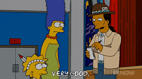The Simpsons gif. Marge Simpson and Lisa Simpson stand in a school theater and look up at a Native American man who is writing on a notepad, saying “Very good.” Lisa wears a feather and beads in her hair and looks up at the man with worry.