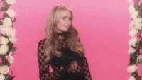 Celebrity gif. Paris Hilton flirtatiously raises a hand and waves at us in front of a pink background flanked by white roses.
