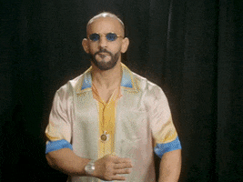 Video gif. Ottman Azaitar wearing a silk shirt and blue tinted glasses strikes a karate pose in front of a black curtain and says, "Come at me bro."