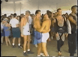 Video gif. Two shirtless men dance near a man in a leotard among other partiers in 1990s attire.