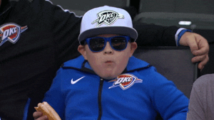 Sports gif. Child wearing OKC jacket, hat, and blue sunglasses in the audience, chewing contentedly on a hot dog.