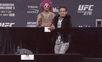 Sports gif. Wearing a furry black jacket and a complicated black-and-white shirt, a very intense Max Holloway claps, then flexes as he yells: Text, "Let's go!" Finally, he slaps his chest and holds up his arms.