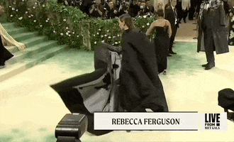 Met Gala 2024 gif. Rebecca Ferguson tosses her Thom Browne black silk moiré cape to one side, showing the full, heavy black fabric gathered around the neckline to great large billowy folds.
