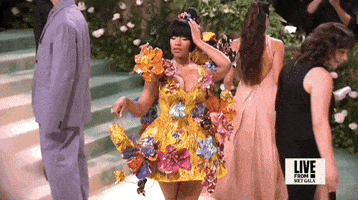 Met Gala 2024 gif. Slow motion shot of Nicki Minaj holding one hand to her head and looking up to her side in a playful pose. She's wearing a yellow Marni mini dress with fringe detailing, a low v-neckline, and 3D multicolored metal flowers sprouting in all directions around the dress. The skirt flares out and comes to her upper thigh as she dangles a tiny matching yellow purse.