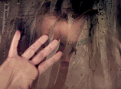 Music video gif. In video for Criminal, Britney Spears stands obscured behind a humid, foggy glass striped with swipe marks, pressing her hand to the glass and looking down.